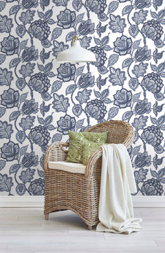 A wicker chair with a white cushion, green patterned pillows, and a cream throw blanket sits in front of Midsummer Jacobean Wallpaper by York Wallcoverings, featuring a dark blue botanical print of flowers and vines.