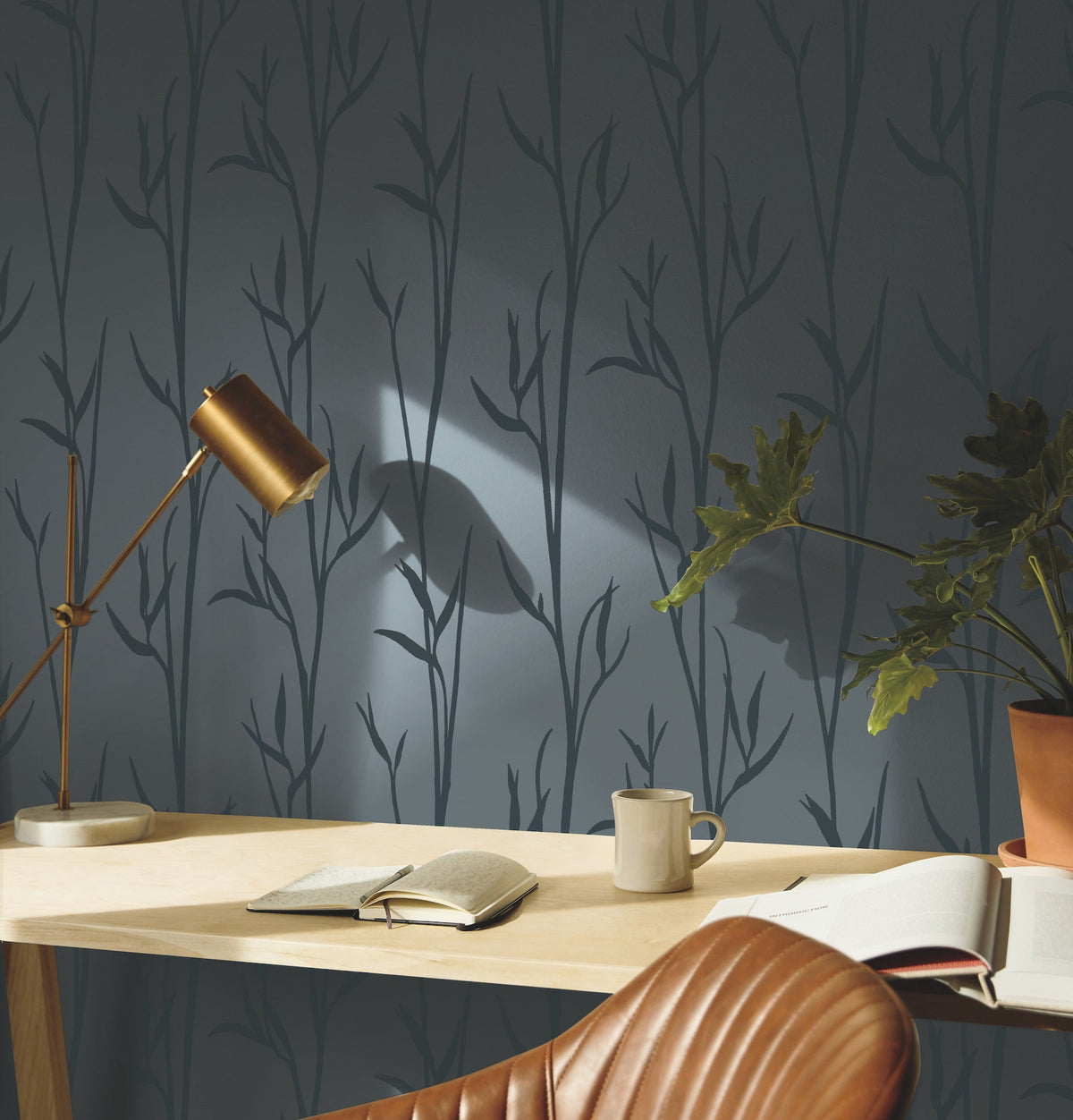 A wooden desk with open books, a mug, a plant, and a gold desk lamp sits in front of a dark wall adorned with York Wallcoverings’ Matcha Wallpaper, featuring a subtle leafy pattern. A brown leather chair is partially visible in the foreground.