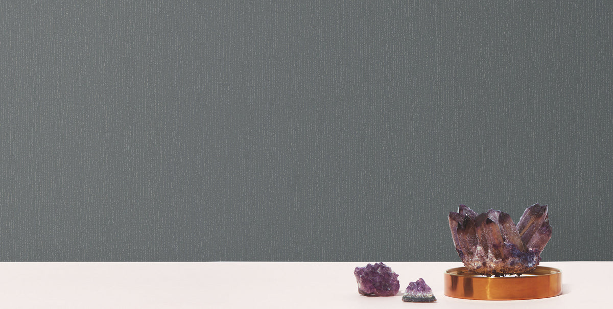 Three amethyst crystals, one large on a gold tray and two smaller nearby, rest on a white surface in front of a backdrop covered with Radiant Juniper Wallpaper by York Wallcoverings.