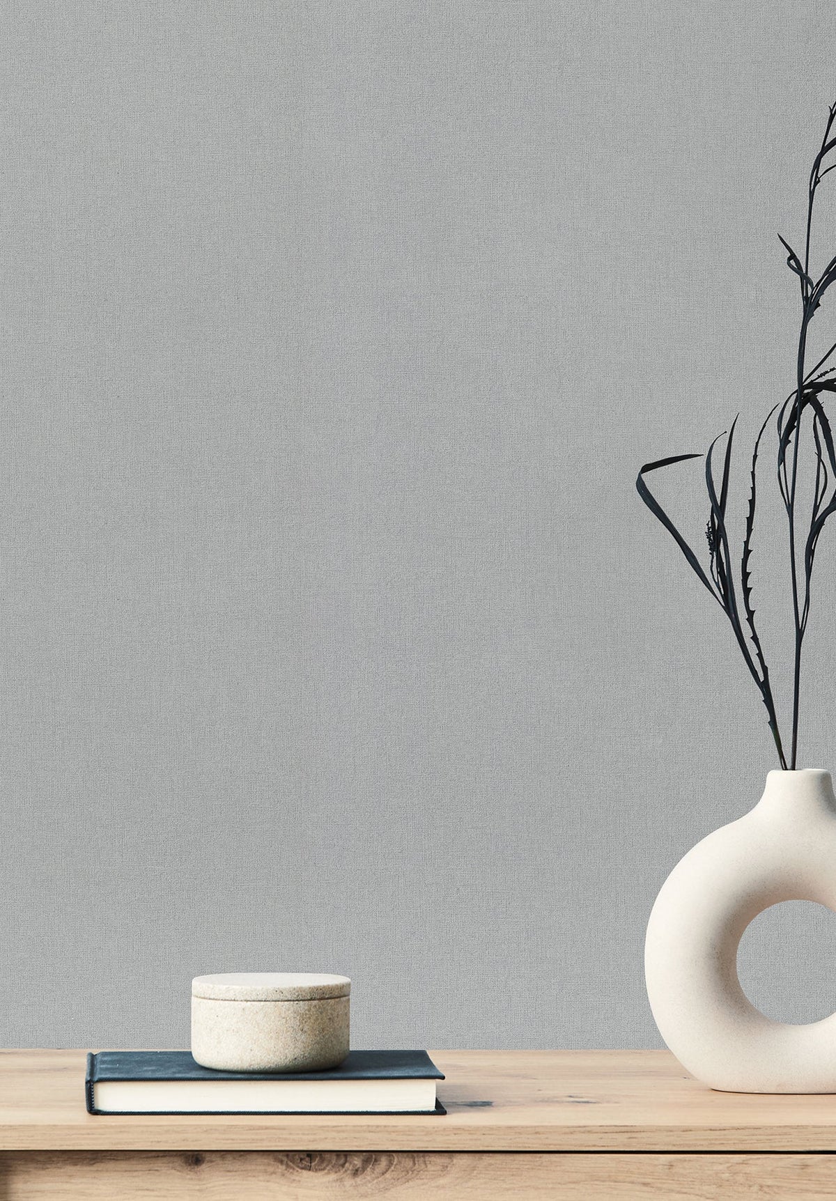 A textured light gray Gypsy Weave Wallpaper serves as the background for a wooden surface featuring a stack of a book and a small container, alongside a round vase containing dried grass.