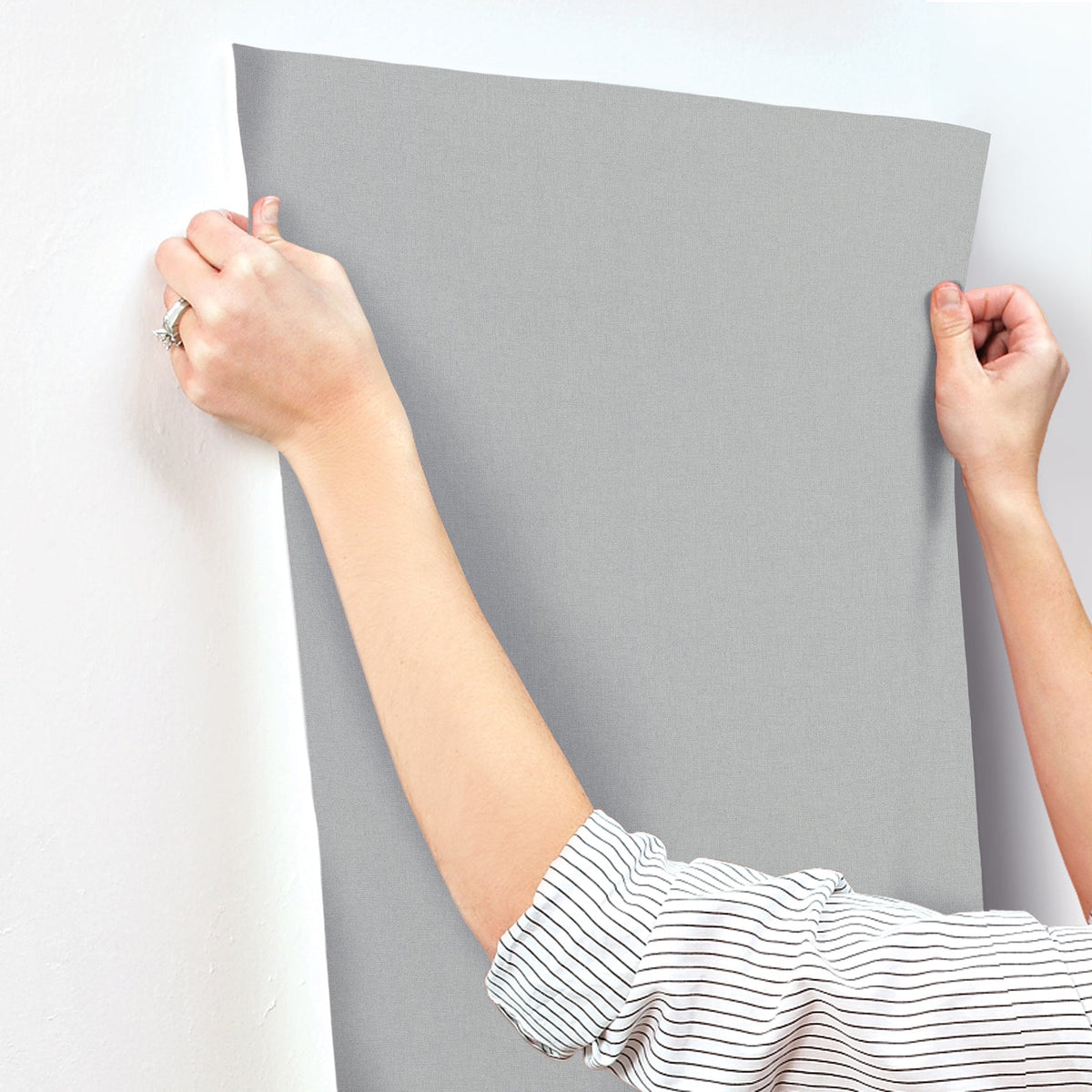 Hands applying a sheet of solid grey Gypsy Weave Wallpaper by York Wallcoverings to a wall.