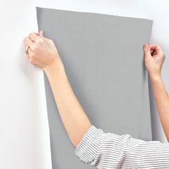 Hands applying a sheet of solid grey Gypsy Weave Wallpaper by York Wallcoverings to a wall.