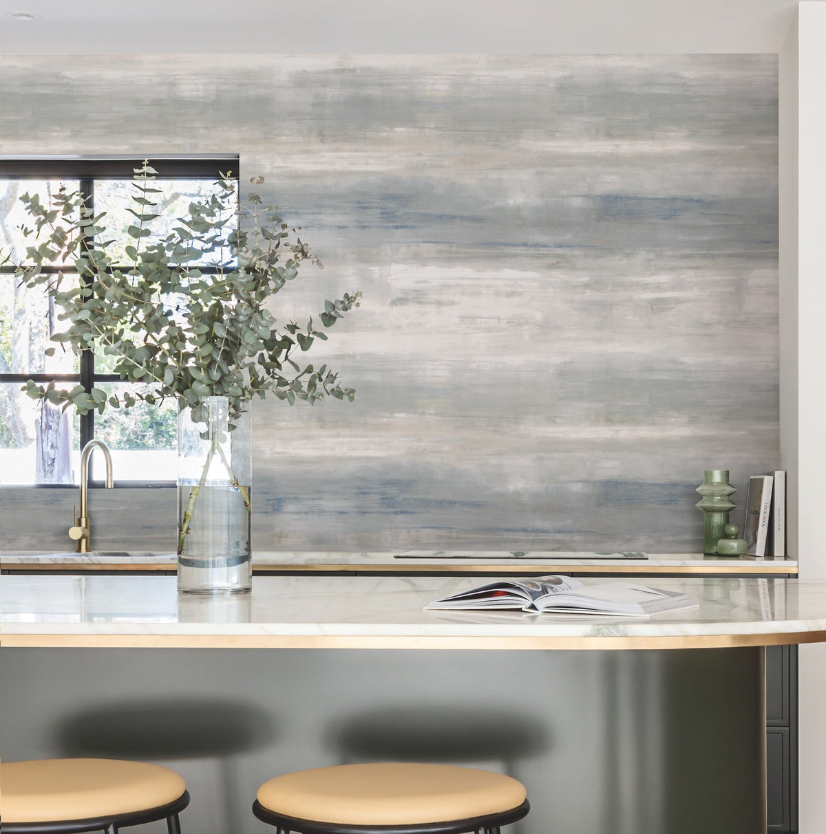 A modern kitchen featuring a marble island, two beige stools, a glass vase with eucalyptus branches, an open book, green candles, and a blue-gray accent wall adorned with York Wallcoverings’ Transition Wallpaper, all illuminated by natural light streaming through the window.