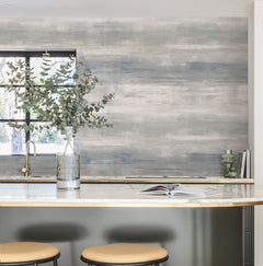 A modern kitchen featuring a marble island, two beige stools, a glass vase with eucalyptus branches, an open book, green candles, and a blue-gray accent wall adorned with York Wallcoverings’ Transition Wallpaper, all illuminated by natural light streaming through the window.