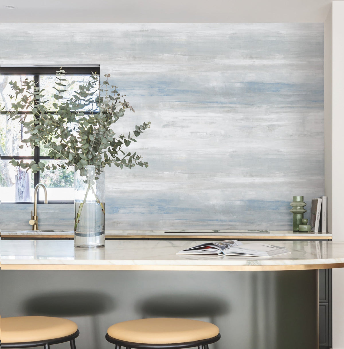 A modern kitchen featuring a marble countertop, two beige stools, a glass vase filled with leafy branches, an open book, and a window that allows natural light to pour in. The wall showcases York Wallcoverings’ Transition Wallpaper in a blue and white textured design.