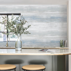 A modern kitchen featuring a marble countertop, two beige stools, a glass vase filled with leafy branches, an open book, and a window that allows natural light to pour in. The wall showcases York Wallcoverings’ Transition Wallpaper in a blue and white textured design.
