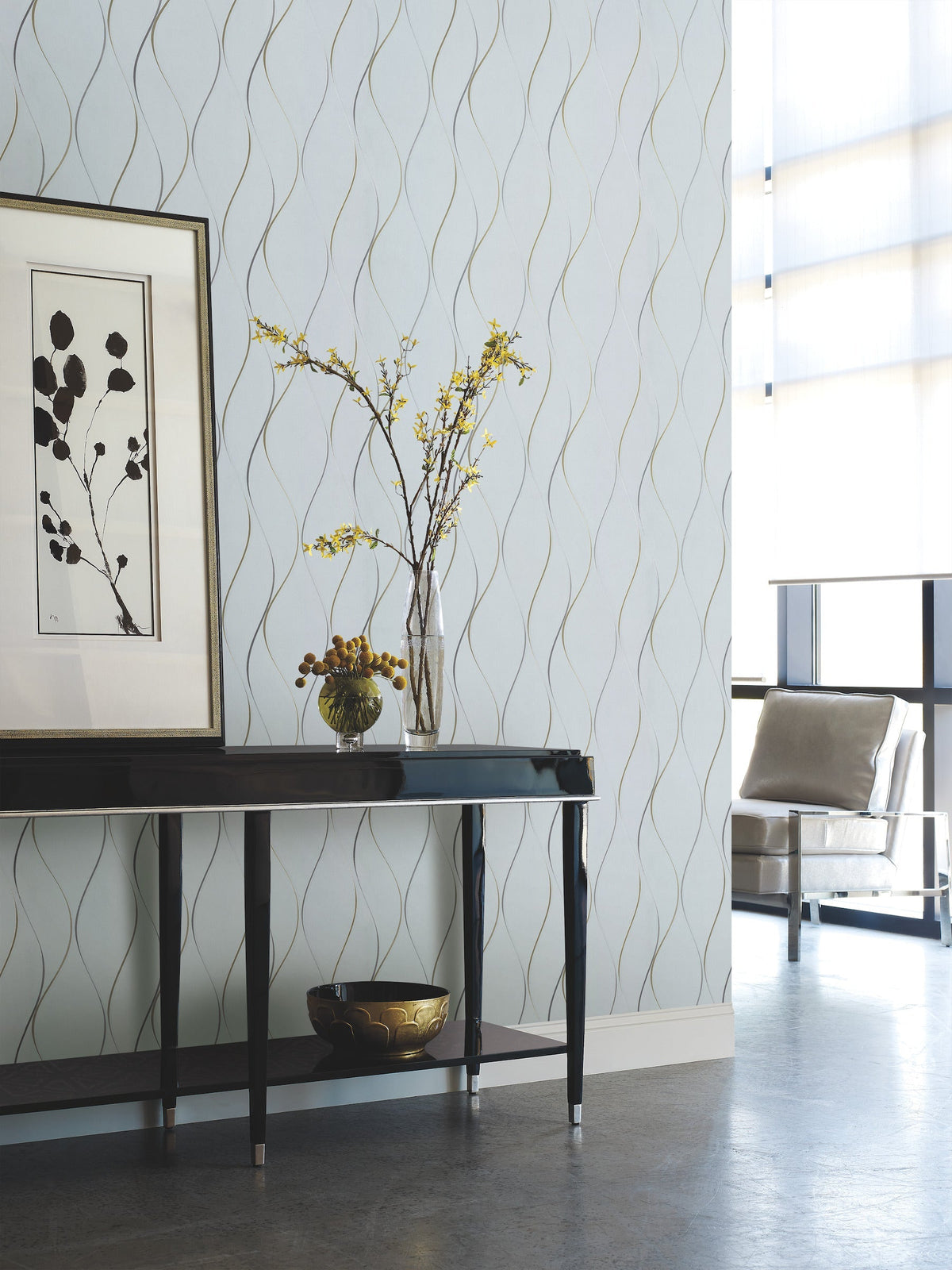 A modern interior featuring York Wallcoverings’ Wavy Stripe Wallpaper, a black console table topped with a vase of yellow flowers, framed art, and a gold bowl. A chair by tall windows completes the minimalist decor.
