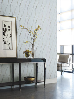A modern interior featuring York Wallcoverings’ Wavy Stripe Wallpaper, a black console table topped with a vase of yellow flowers, framed art, and a gold bowl. A chair by tall windows completes the minimalist decor.