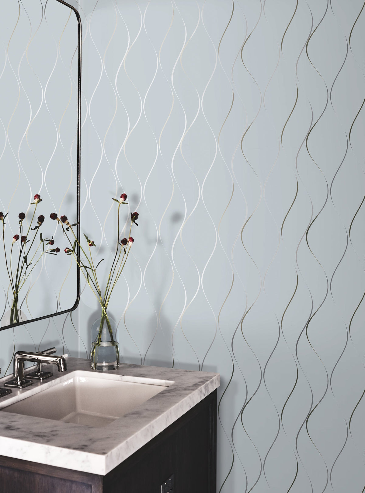 A bathroom corner features a white sink with a marble countertop, a modern faucet, and a glass vase displaying red and black flowers. The wall is decorated with York Wallcoverings’ Wavy Stripe Wallpaper in light blue, showcasing a wavy metallic line pattern.