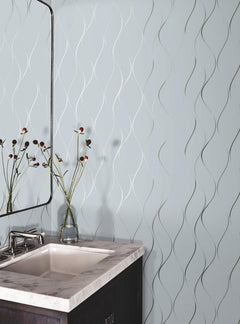 A bathroom corner features a white sink with a marble countertop, a modern faucet, and a glass vase displaying red and black flowers. The wall is decorated with York Wallcoverings’ Wavy Stripe Wallpaper in light blue, showcasing a wavy metallic line pattern.