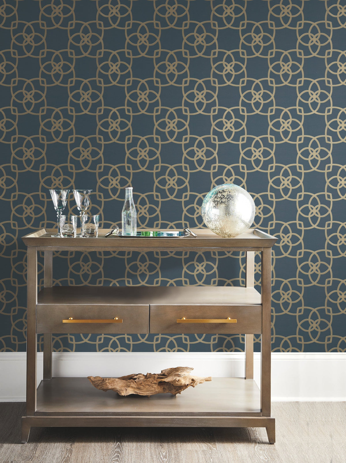 A wooden bar cart with glassware, bottles, a globe, and driftwood sits in front of a dark blue wall adorned with York Wallcoverings’ Serendipity Wallpaper.