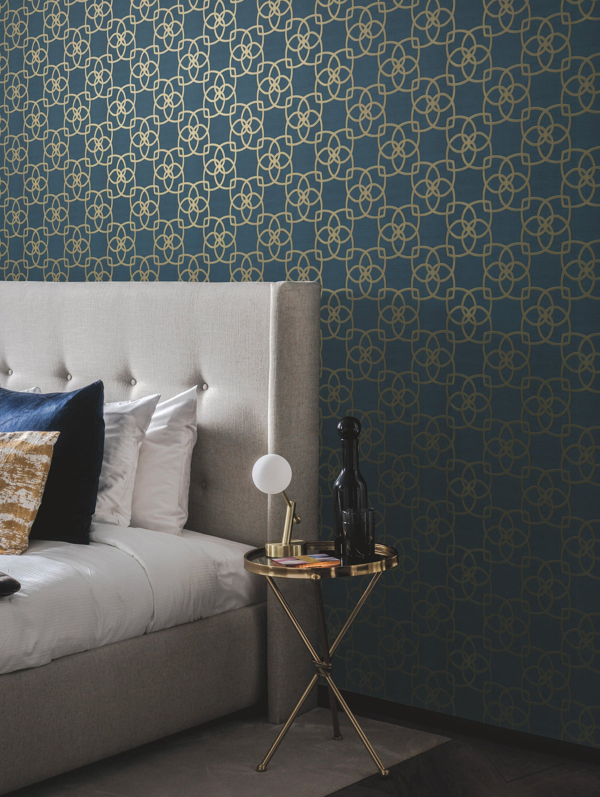 A modern bedroom featuring a tufted gray headboard, white bedding, a blue and gold pillow, and a small round gold side table holding a lamp, a black vase, and a glass. The wall is adorned with the “Serendipity Wallpaper” by York Wallcoverings in a blue and gold geometric pattern.