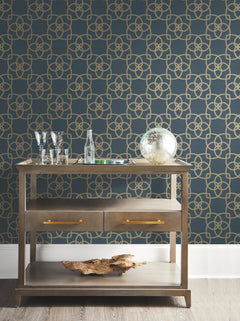 A wooden console table adorned with glassware, a glass bottle, a globe, and decorative accents stands against a blue wall featuring the Serendipity Wallpaper by York Wallcoverings, showcasing a striking gold geometric pattern. The table includes two drawers and a lower shelf displaying a piece of driftwood.
