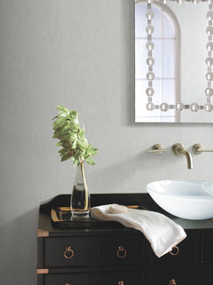 A modern bathroom featuring York Wallcoverings’ Natural Texture Wallpaper, a black vanity, glass vessel sink, wall-mounted faucet, towel, and a glass vase holding green leaves. A decorative mirror reflects a window in the background.