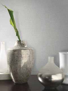 A speckled ceramic vase with a single green leaf stands beside a round, metallic vase on a dark surface, with two white vases in the background set against York Wallcoverings’ Natural Texture Wallpaper in light gray.