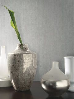 A tall ceramic vase with a green leaf stands next to a smaller, rounded metallic vase on a dark surface. Two white vases are blurred in the background against the light gray Natural Texture Wallpaper by York Wallcoverings.