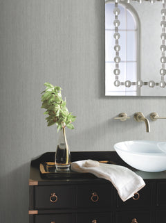 A modern bathroom features Natural Texture Wallpaper by York Wallcoverings as a backdrop, complemented by a black vanity with brass handles, a clear glass vase with green leaves, a folded white towel, a white basin sink, wall-mounted faucets, and a decorative mirror reflecting a window.