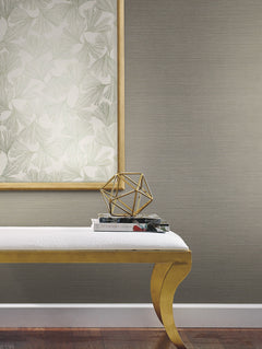 A white bench with gold legs sits on a wood floor, holding two books and a gold geometric sculpture. Behind it, a large framed botanical print hangs on a wall covered in York Wallcoverings’ Shining Sisal Grasscloth Wallpaper.