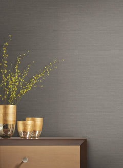 Shining Sisal Grasscloth Wallpaper in dark grey with a fine horizontal texture, shown behind gold vases and a branch with small yellow flowers on a wooden surface.