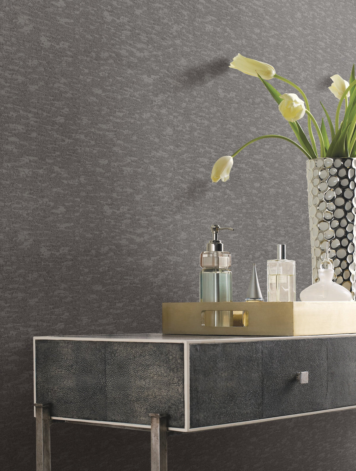 A modern table with a metallic frame holds a tray with perfume bottles, soap dispenser, and a vase of drooping white tulips, set against a wall covered in Weathered Cypress Wallpaper by York Wallcoverings.