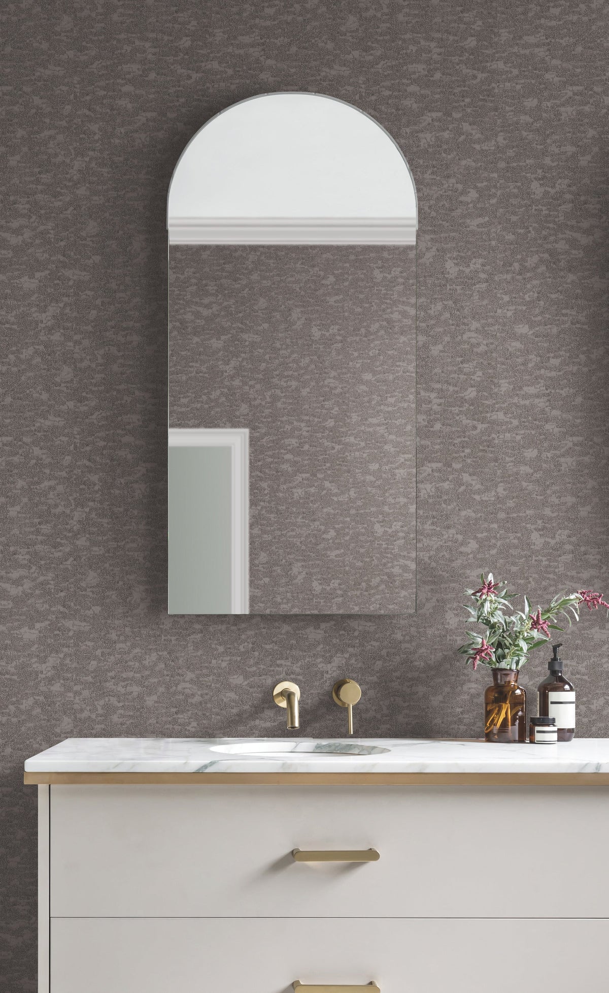 A bathroom vanity with a marble countertop, gold faucet, and an arched mirror stands against a Weathered Cypress Wallpaper by York Wallcoverings. A small vase with flowers and a soap dispenser sit on the counter in front of the textured gray design.