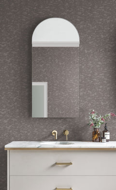 A modern bathroom features a marble countertop, gold faucet, and a rectangular mirror with an arched top. A small vase of flowers and toiletries sit on the sink, all set against York Wallcoverings’ Weathered Cypress Wallpaper for a textured gray backdrop.