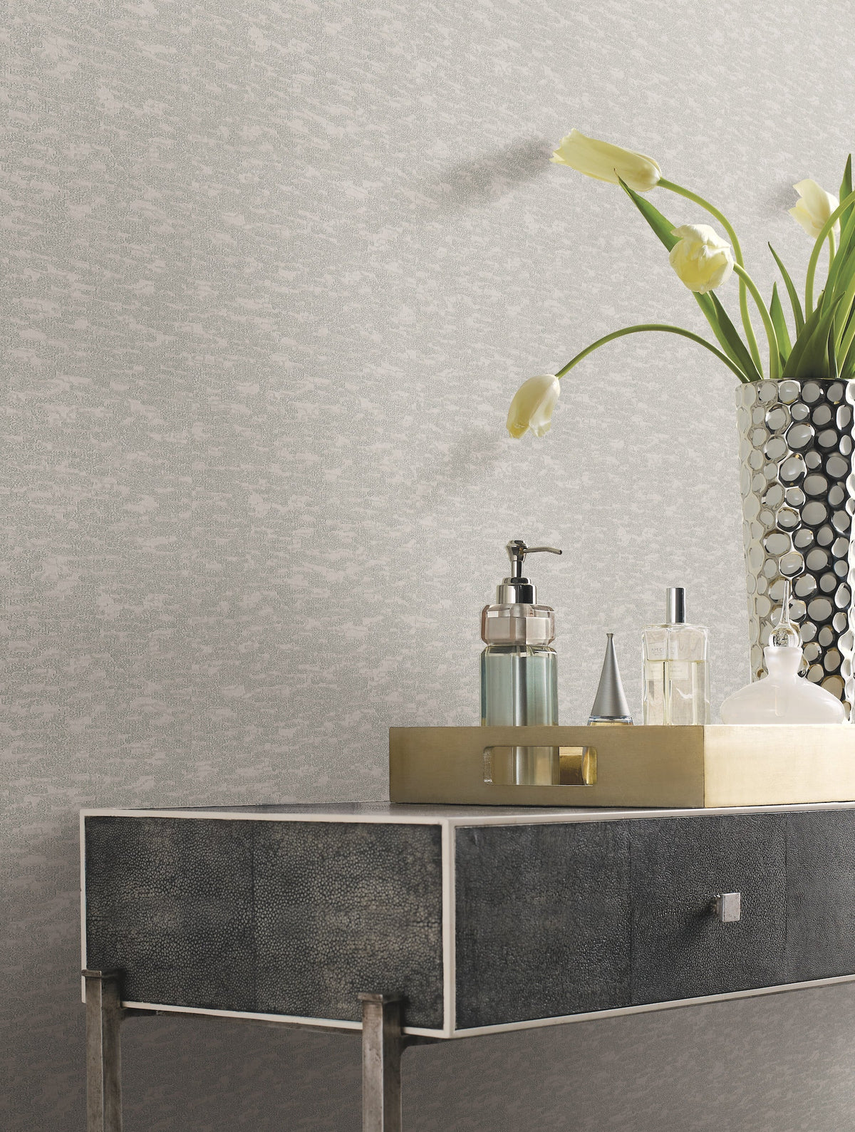 A metallic tray with toiletries and a faceted vase holding white tulips sits on a modern, dark-textured console table against a wall adorned with York Wallcoverings’ Weathered Cypress Wallpaper.