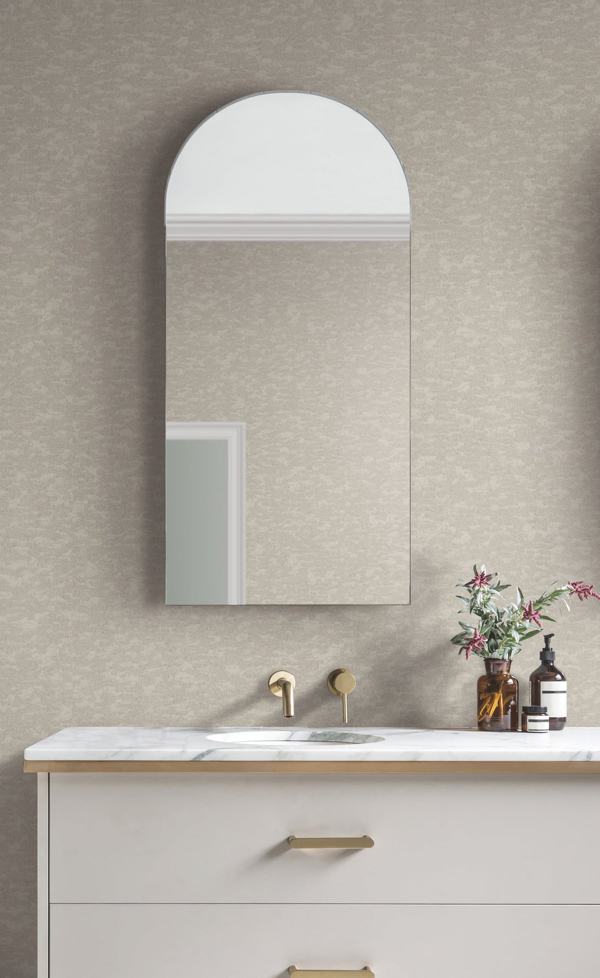 A modern bathroom features a marble countertop, gold faucet, and arched mirror. A small vase with flowers and a soap dispenser sit on the counter. The wall is covered in Weathered Cypress Wallpaper by York Wallcoverings.
