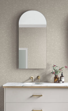 A modern bathroom features a marble countertop, gold faucet, and arched mirror. A small vase with flowers and a soap dispenser sit on the counter. The wall is covered in Weathered Cypress Wallpaper by York Wallcoverings.