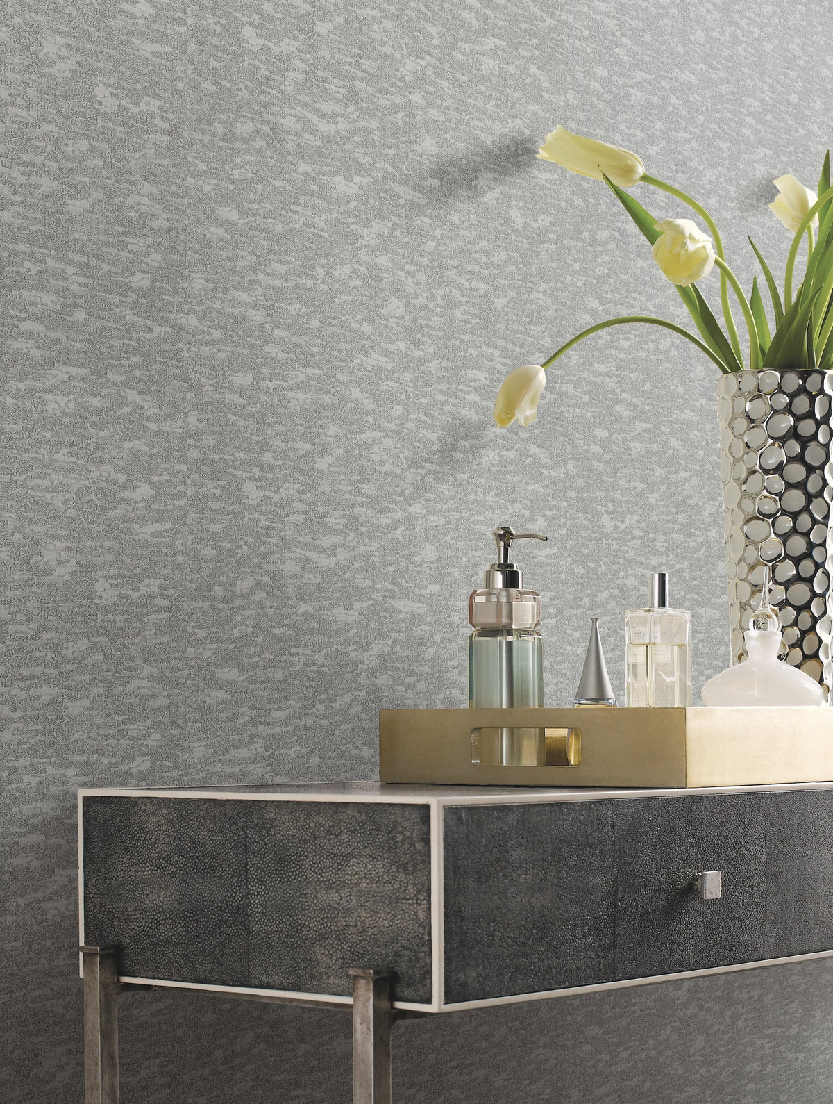 A modern console table with a metallic tray holding soap, lotion, and perfume bottles stands beside a vase of yellow tulips set against York Wallcoverings’ Weathered Cypress Wallpaper.