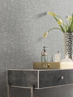 A modern console table with a metallic tray holding soap, lotion, and perfume bottles stands beside a vase of yellow tulips set against York Wallcoverings’ Weathered Cypress Wallpaper.