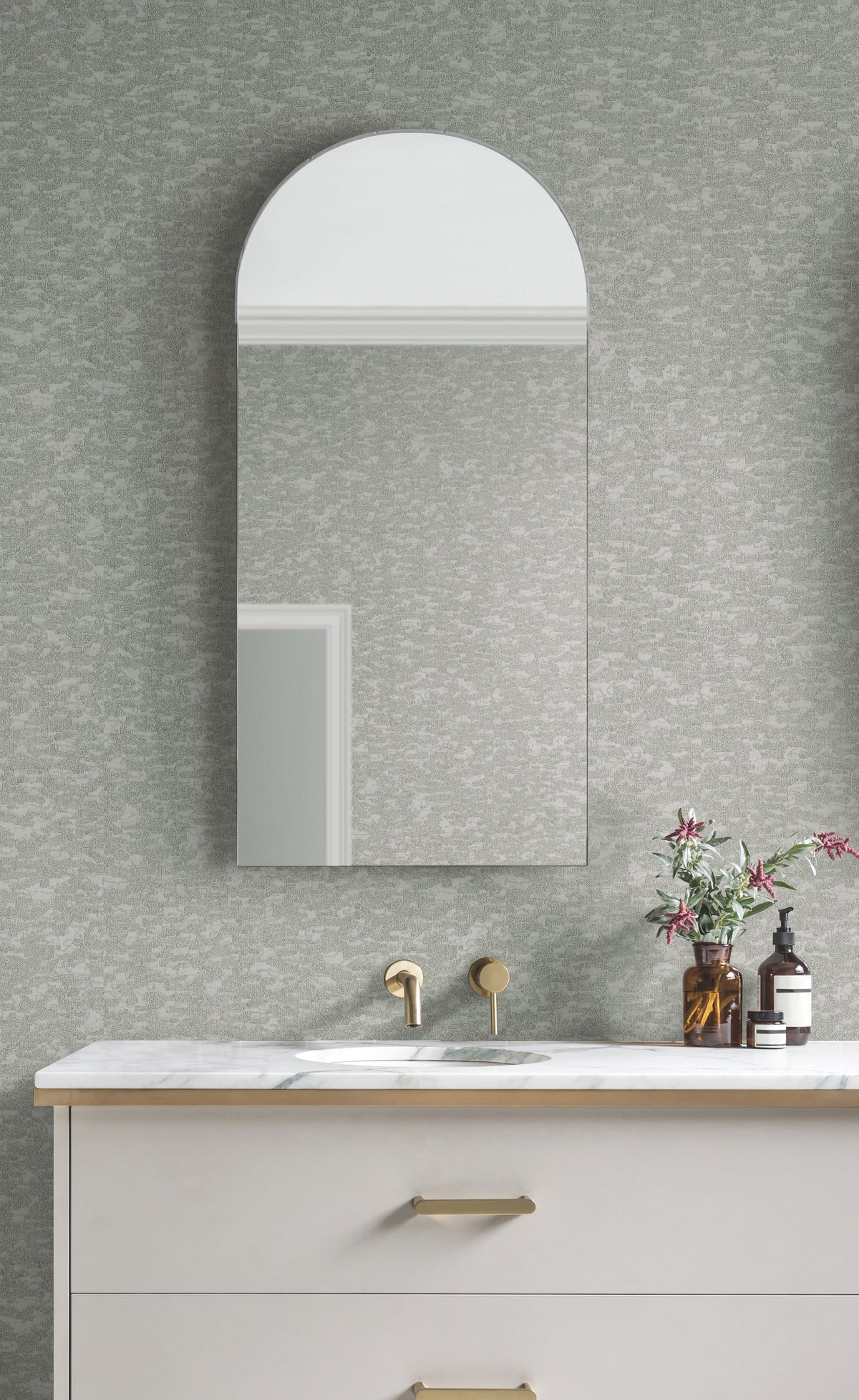 A modern bathroom features a marble sink, gold faucet, arched mirror, and flowers in a vase. The wall is adorned with York Wallcoverings’ Weathered Cypress Wallpaper in a textured gray pattern, while a bottle of soap sits on the counter.