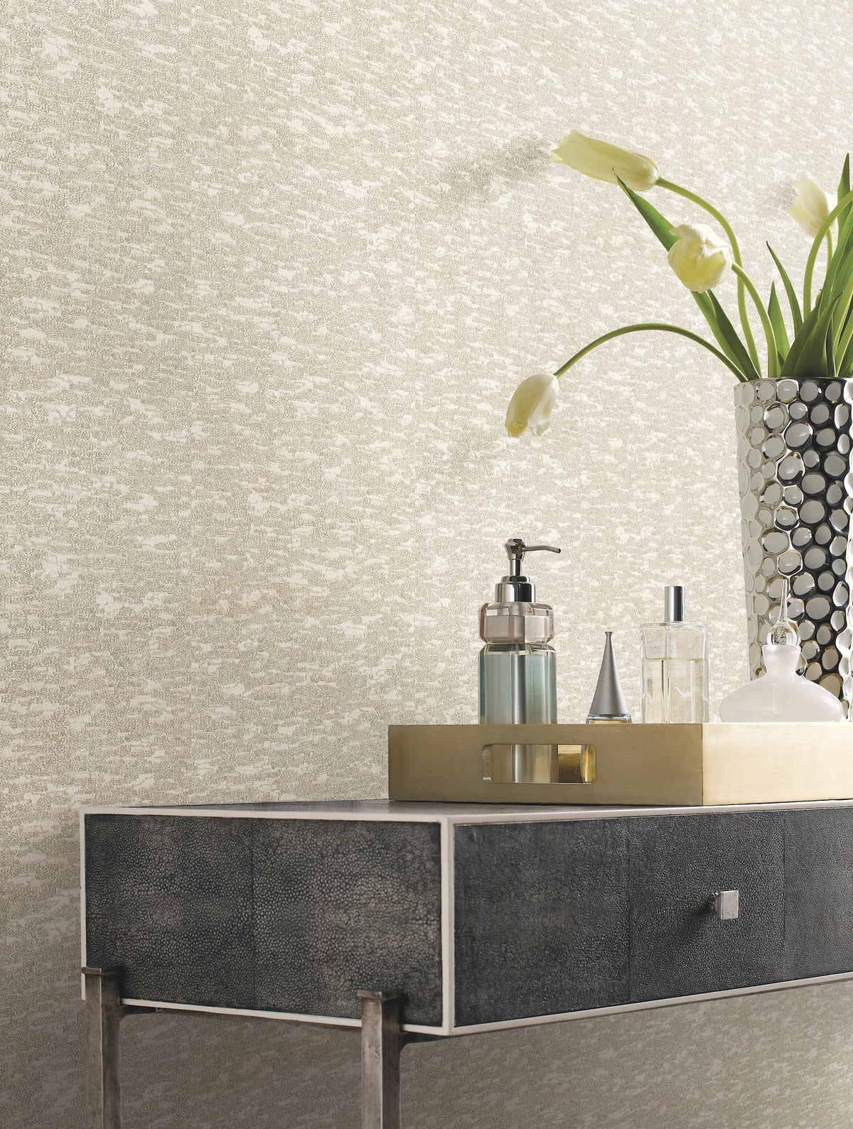 A metallic tray with bottles and a soap dispenser sits on a dark textured console table; a tall silver vase filled with white tulips stands beside it, set against York Wallcoverings’ Weathered Cypress Wallpaper.