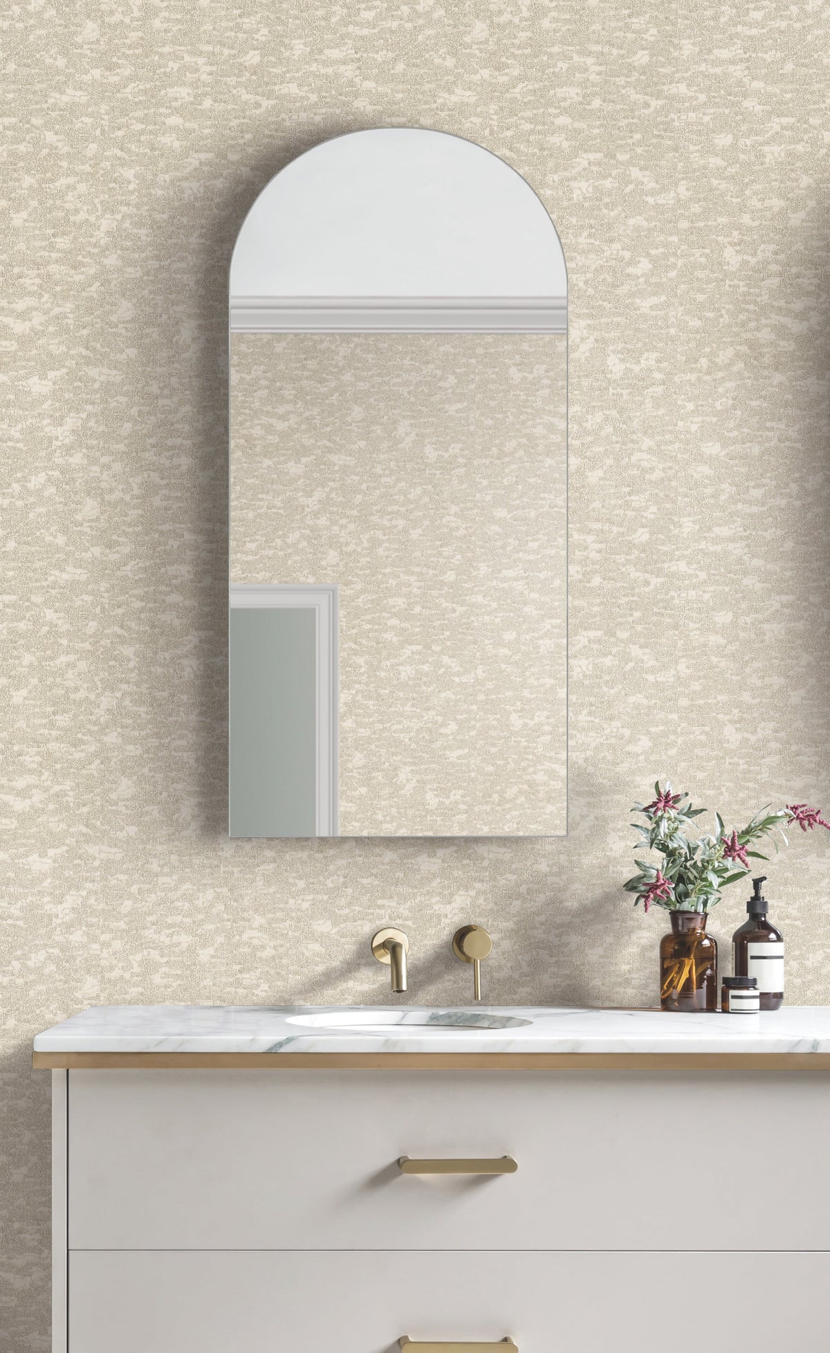 A modern bathroom features a rounded rectangular mirror above a white marble sink with gold fixtures, set against walls adorned in Weathered Cypress Wallpaper by York Wallcoverings. A vase with flowers and a soap dispenser sit on the countertop.