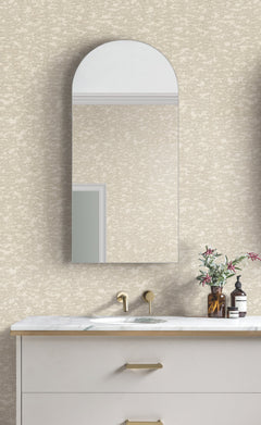 A modern bathroom features a rounded rectangular mirror above a white marble sink with gold fixtures, set against walls adorned in Weathered Cypress Wallpaper by York Wallcoverings. A vase with flowers and a soap dispenser sit on the countertop.