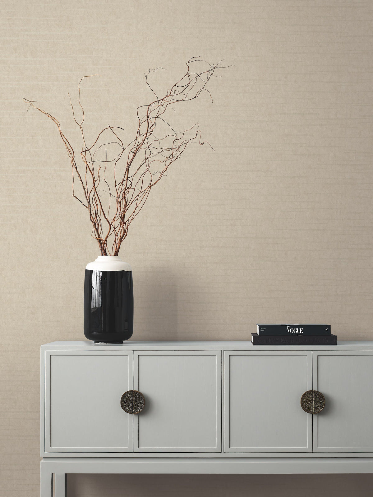 A minimalist console table with two drawers, a black vase holding bare branches, and two stacked magazines sits against a beige Tiger’s Eye Wallpaper by York Wallcoverings.