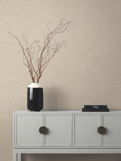 A minimalist console table with two drawers, a black vase holding bare branches, and two stacked magazines sits against a beige Tiger’s Eye Wallpaper by York Wallcoverings.