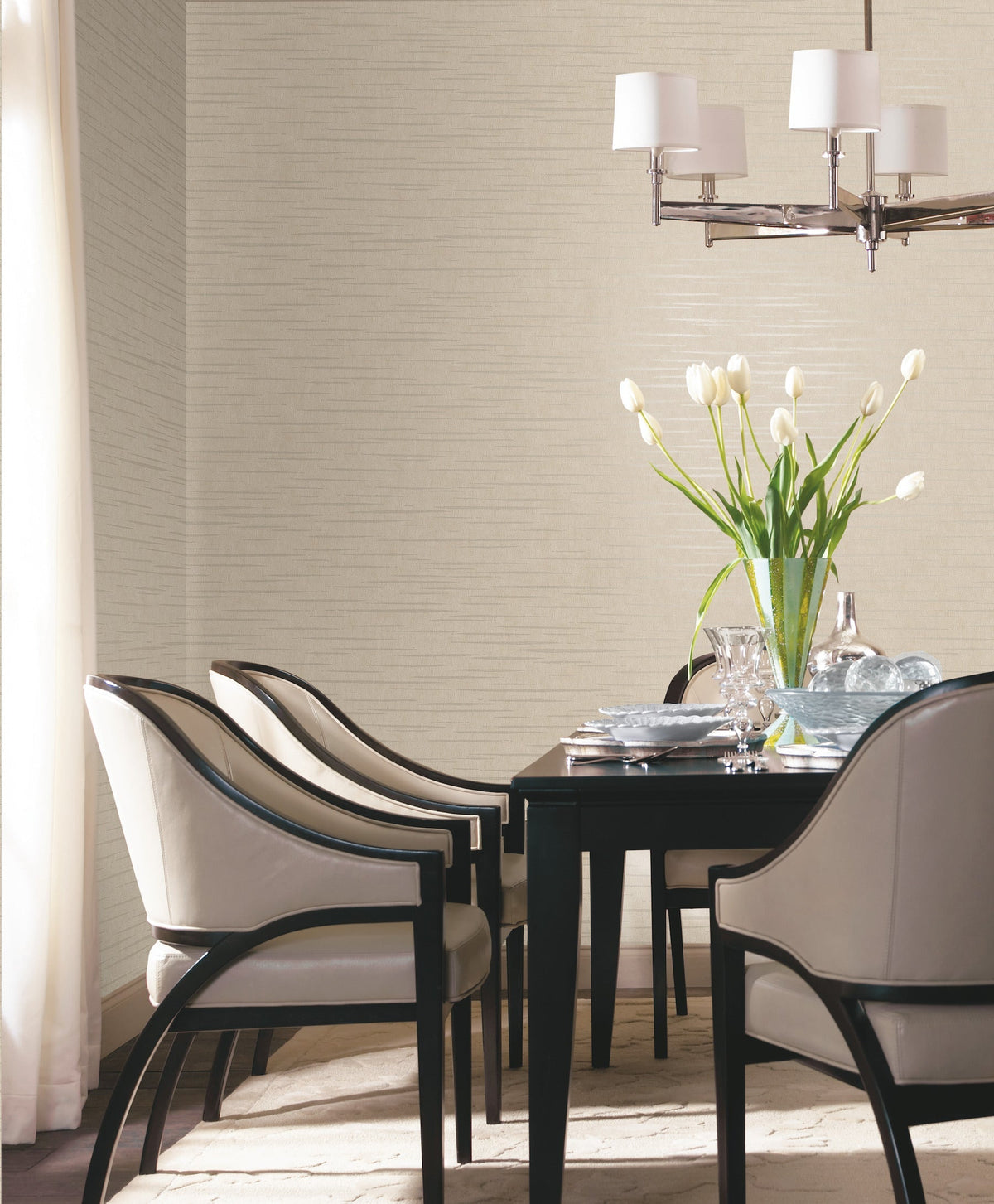 A modern dining room featuring Tiger’s Eye Wallpaper by York Wallcoverings on the walls, a dark table set for four, cream upholstered chairs, a vase of white tulips, and a chrome chandelier with white shades. Sunlight shines through a curtain on the left.