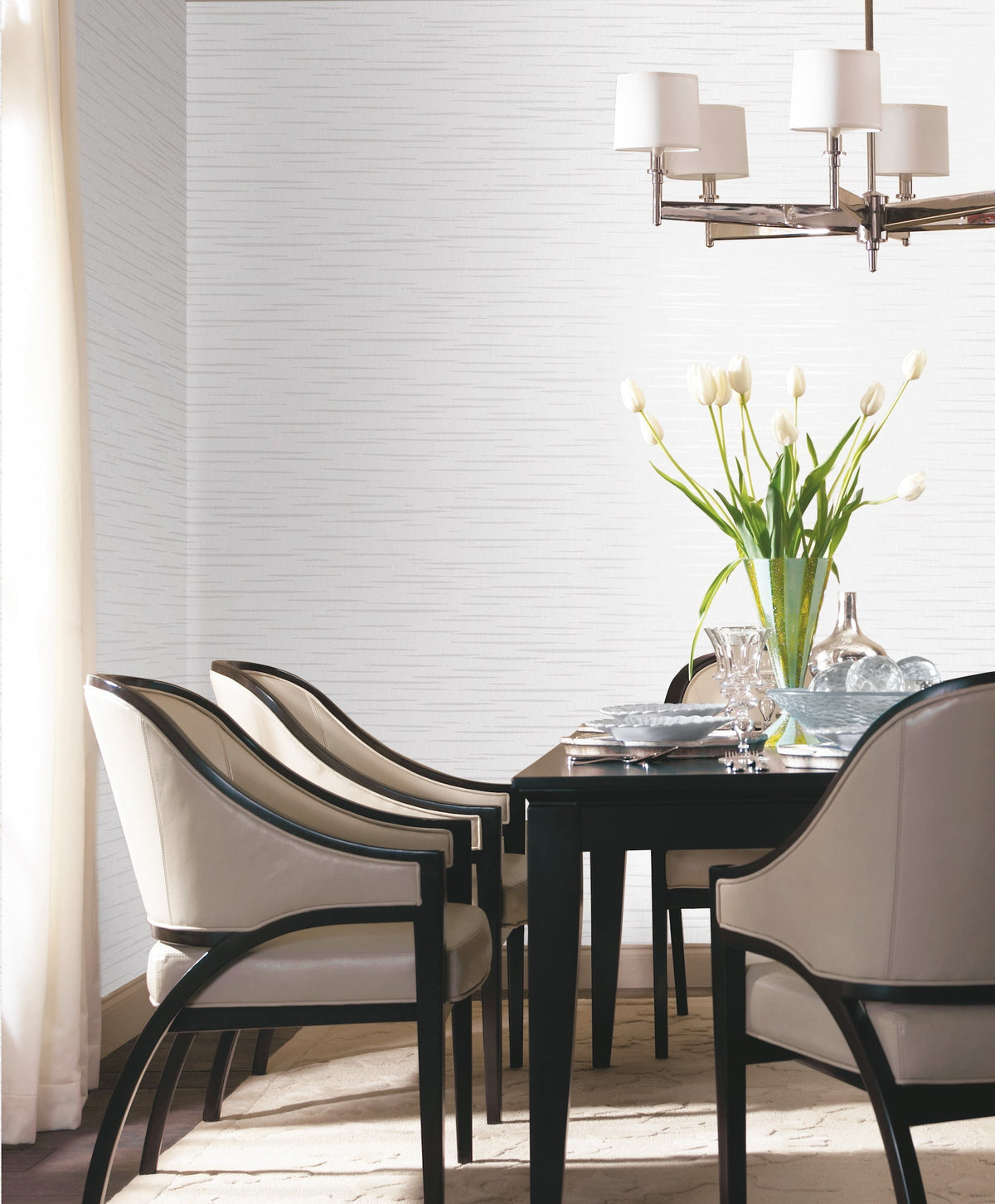 A modern dining room featuring York Wallcoverings’ Tiger’s Eye Wallpaper, a dark table with four cushioned chairs, a vase of white tulips, clear glassware, and a silver chandelier. Light pours in from a window with sheer curtains.