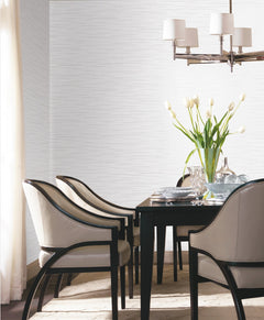 A modern dining room featuring York Wallcoverings’ Tiger’s Eye Wallpaper, a dark table with four cushioned chairs, a vase of white tulips, clear glassware, and a silver chandelier. Light pours in from a window with sheer curtains.