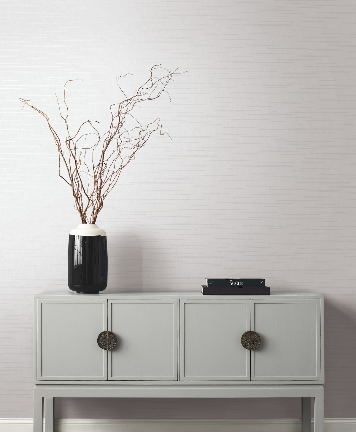 A minimalist gray console table with two large circular knobs holds a black vase with tall, bare branches and a small stack of black books, set against a light, textured wall featuring the Tiger’s Eye Wallpaper by York Wallcoverings.