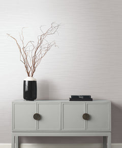 A minimalist gray console table with two large circular knobs holds a black vase with tall, bare branches and a small stack of black books, set against a light, textured wall featuring the Tiger’s Eye Wallpaper by York Wallcoverings.