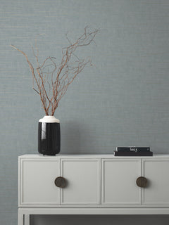 A minimalist sideboard with round handles holds a tall black vase with bare branches and a small stack of black books, set against York Wallcoverings’ Tiger’s Eye Wallpaper in light gray.