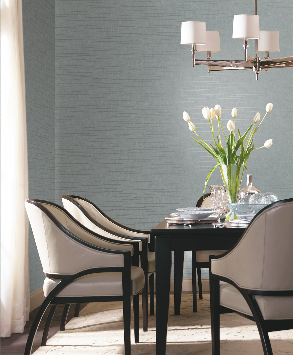 Modern dining room featuring York Wallcoverings’ Tiger’s Eye Wallpaper as a backdrop, with a black table, four beige chairs, a vase of white tulips, and a chandelier overhead. Light filters in through white curtains, creating a bright, elegant atmosphere.
