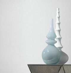 Two decorative vases sit on a glass table against a wall adorned with Gossamer Woven Wallpaper by York Wallcoverings. One vase is light blue with a curvy shape, and the other is white with a segmented, stacked design.