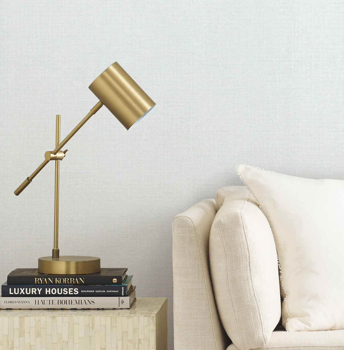 A gold desk lamp sits on a small table stacked with books next to a beige sofa with a white pillow, set against a light textured wall featuring the Gossamer Woven Wallpaper by York Wallcoverings.