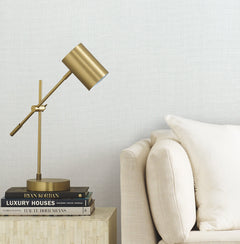 A gold desk lamp sits on a small table stacked with books next to a beige sofa with a cream pillow, set against a light textured wall covered in Gossamer Woven Wallpaper by York Wallcoverings.
