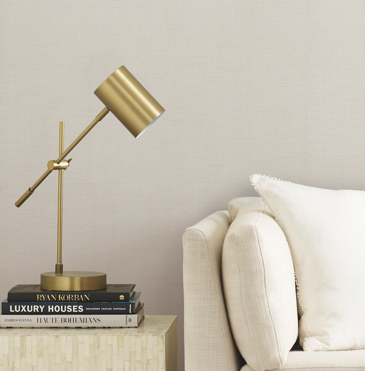 A brass desk lamp sits on a stack of books beside a beige sofa with a white pillow, against a wall adorned with York Wallcoverings’ Gossamer Woven Wallpaper.