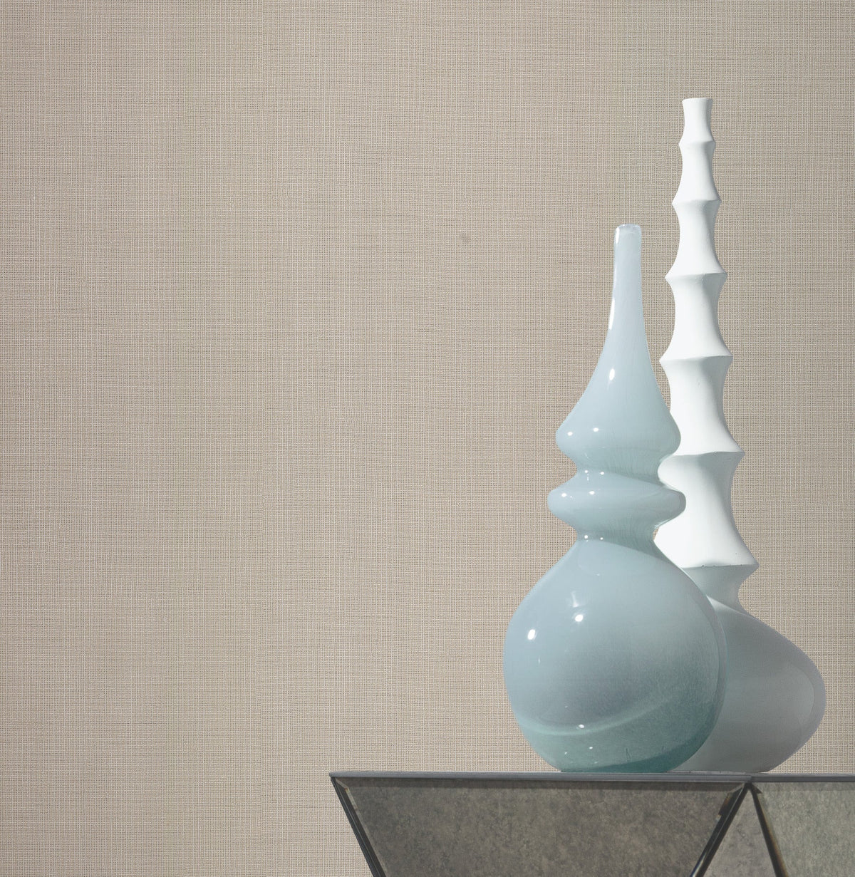 Two modern vases, one glossy light blue with curved shapes and one matte white with stacked segments, stand on a glass surface against a wall adorned with Gossamer Woven Wallpaper by York Wallcoverings.