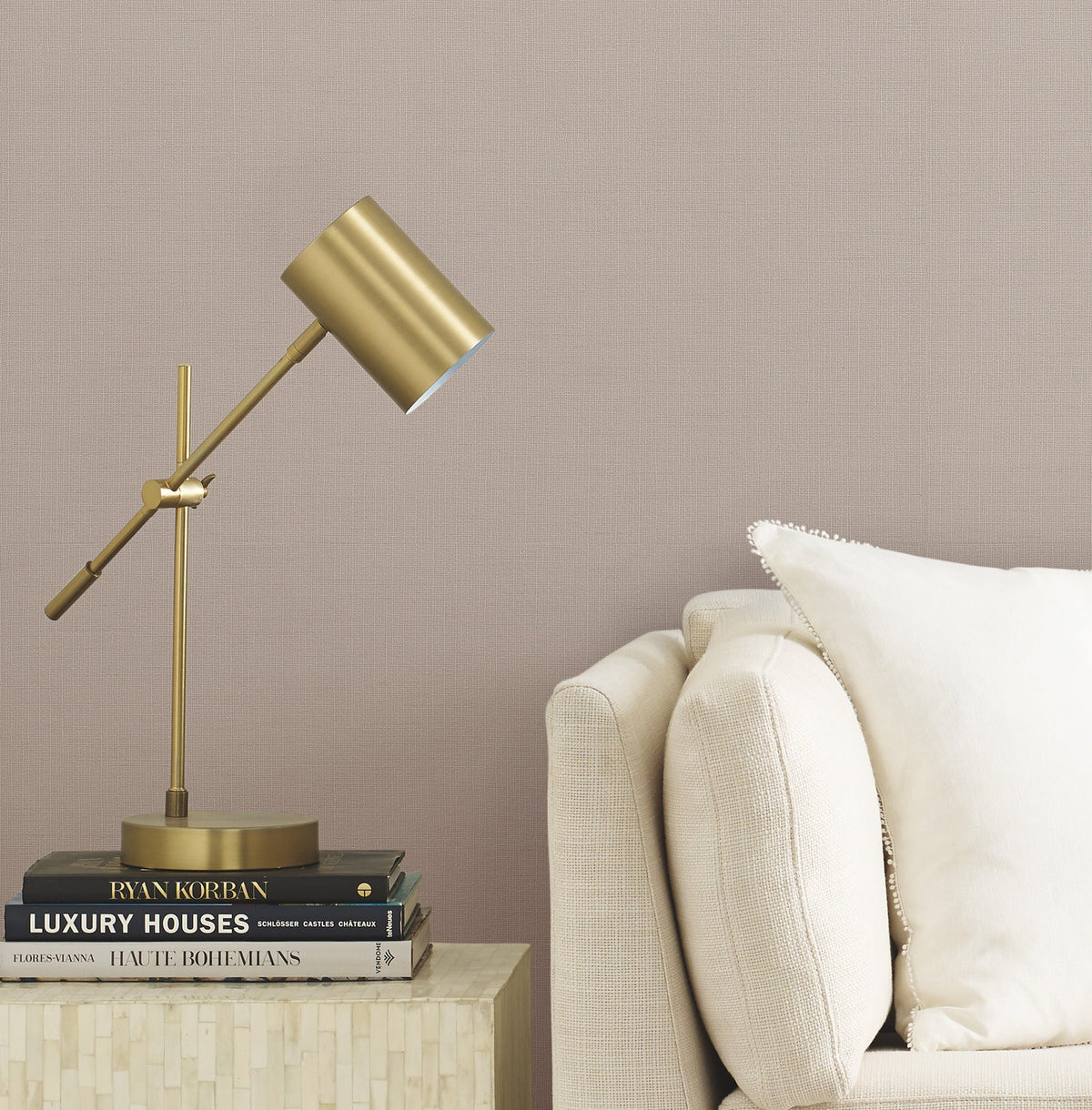 A modern brass desk lamp sits on a small table next to a beige sofa with a white pillow. Several stacked books are under the lamp. The background features Gossamer Woven Wallpaper by York Wallcoverings.